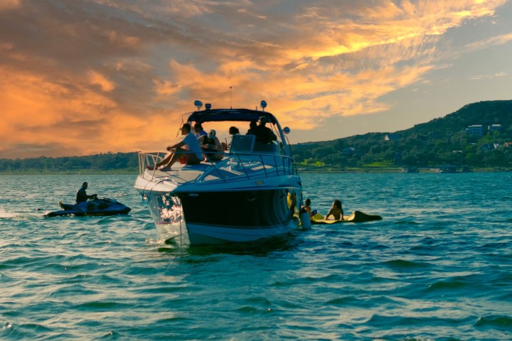 Boat with people on a lake at sunset, with jet ski nearby and hills in background.