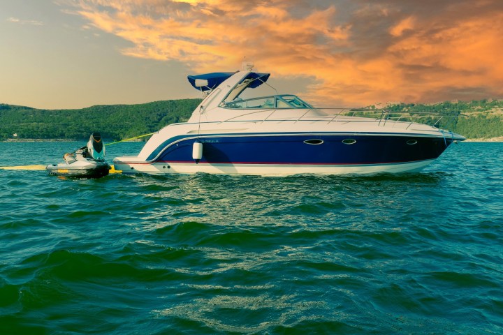 A blue and white yacht on a lake with a jet ski attached, set against an orange sky.