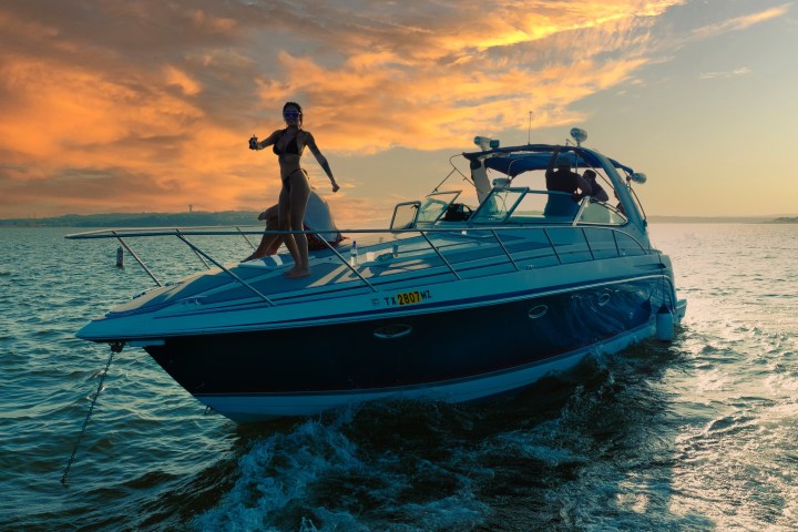 Woman on a yacht at sunset with vibrant orange and blue sky, ocean waves behind.